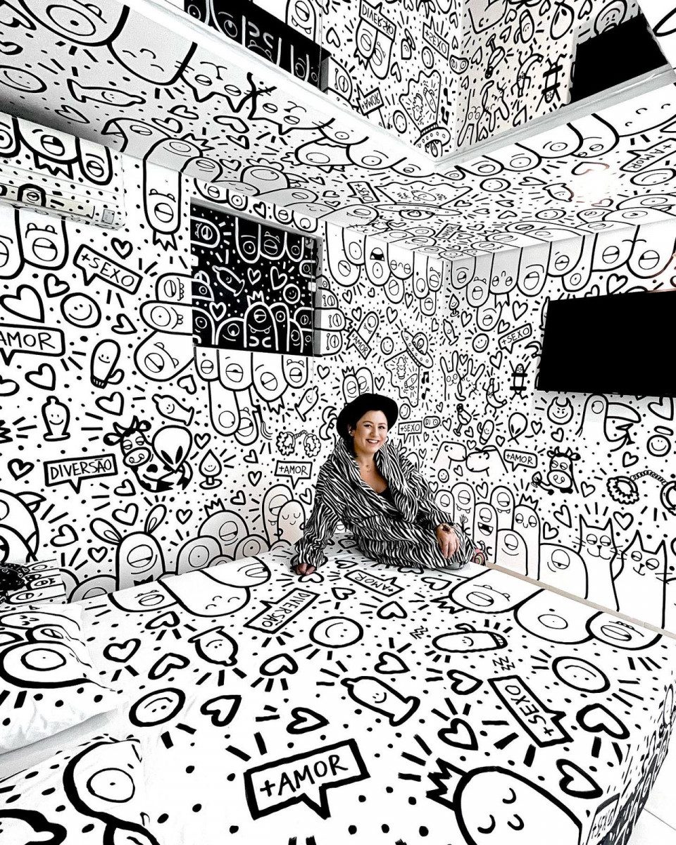 Ana sentada na cama da suíte branca com ilustrações em preto